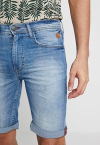 Ljusblå denimshorts med uppvikta fållar, med framfickor och en brun läderlapp på midjebandet. Mönstrad skjorta synlig ovanför.