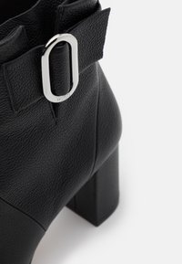 Bottine en cuir noir avec une finition texturée, talon carré et une boucle ovale argentée en accent à la cheville.