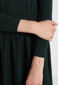 Mörkgrön stickad klänning med figursydd liv och utsvängt kjol. Långa ärmar med ribbade muddar. Tyget verkar mjukt och strukturerat.