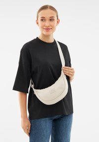 Borsa a tracolla a mezzaluna beige realizzata in morbido tessuto con una struttura imbottita, accompagnata da una tracolla leggera e un piccolo etichetta con il marchio.