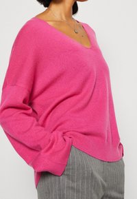 Suéter rosa de cuello en V con mangas largas, hecho de un material suave, con un corte holgado y textura acanalada, combinado con pantalones rayados grises.