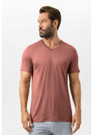 Mann mit braunen Haaren und Bart, der ein staubrosa V-Ausschnitt-T-Shirt und gestreifte Hosen trägt, blickt nach links vor einem weißen Hintergrund.