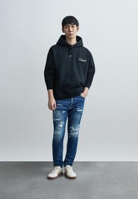 Must kapuutsiga dressipluus valge logoga, kombineeritud kulunud siniste teksade ja beežide tossudega, mille taldadel on pruunid detailid.