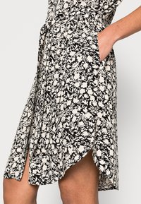 Abito floreale nero e crema con un design fluido, tasche laterali e un orlo asimmetrico con una vita leggermente arricciata.