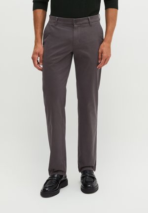 Mann trägt dunkelgraue maßgeschneiderte Hose mit schwarzen Leder-Loafers und einem schwarzen Langarmhemd und steht vor einem schlichten Hintergrund.