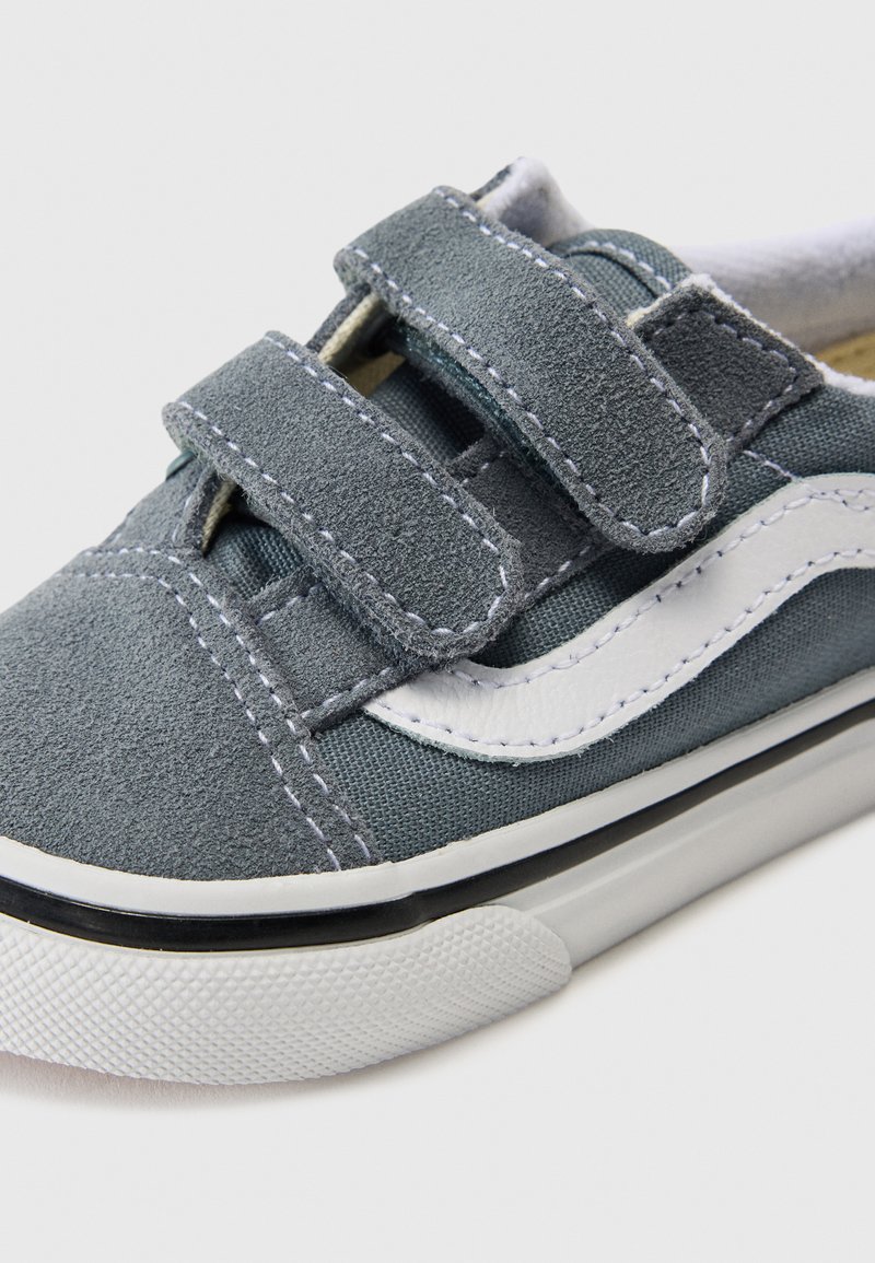 Grijs suède sneaker met witte accenten, voorzien van twee klittenbandsluitingen, gestructureerde stof en een rubberen zool. Close-up weergave benadrukt de stiksel details.