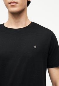 Νεαρό άτομο που φοράει απλό μαύρο κοντομάνικο μπλουζάκι με στρογγυλή λαιμόκοψη και ένα μικρό λευκό κεντητό γράμμα "R" στο αριστερό στήθος.