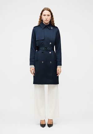 Woman wearing a navy belted trench coat, white wide-leg pants, gray sweater cuffs, and black pointed-toe heels, standing against a plain background.