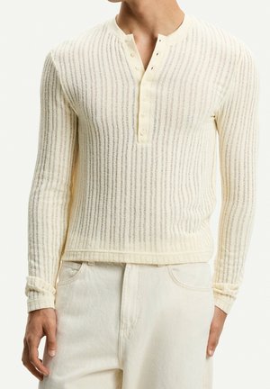 Mann trägt cremefarbenes langärmeliges geripptes Henley-Shirt mit Knopfleiste und offwhite-farbene Hose, gezeigt vom Hals bis zu den Hüften.