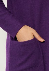 Tessuto di maglione viola che mostra un primo piano della manica e della tasca. Il tessuto appare morbido con una texture liscia e dettagli di cucitura curati.
