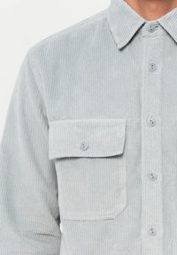 Light gray corduroy shirt with ribbed texture, button closures, and a chest pocket detail. Features a standard collar and visible stitching.