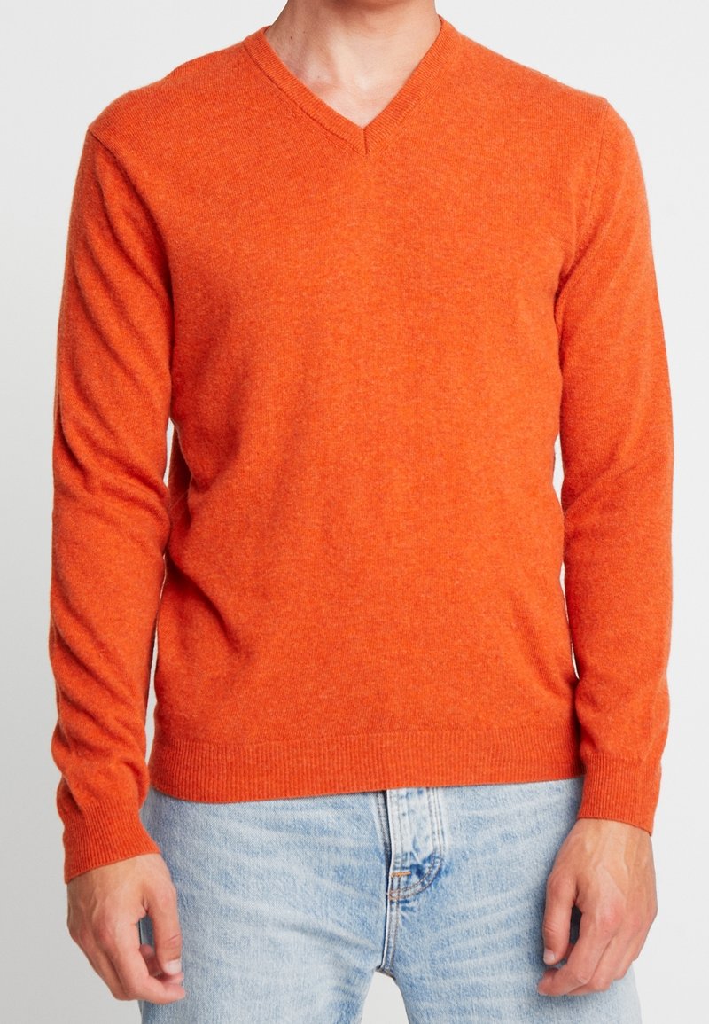 V-halsad orange tröja med långa ärmar, ribbade muddar och nederkant, tillverkad av mjukt material, i kombination med ljusblå denimjeans.