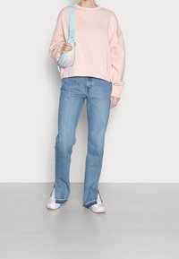 Ljusrosa sweatshirt med rund halsringning och vit logotyp, kombinerad med ljusblå raka jeans och vita sneakers med rosa detaljer.