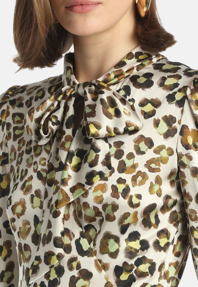 Bluse mit Leopardenmuster aus seidigem Stoff, mit einer geknoteten Schleife am Hals. Besondere Merkmale sind kräftige Brauntöne, Grün und Schwarz auf einem cremfarbenen Hintergrund.