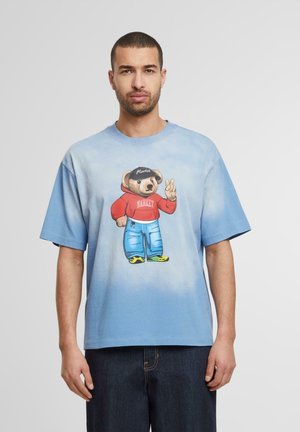 Blaues T-Shirt mit Farbverlauf und Oversized-Passform, mit einem bärentiefer in rotem Kapuzenpullover und blauen Jeans, akzentuiert durch grüne und schwarze Schuhe.