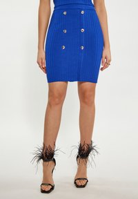 Jupe mini bleue à côtes avec des boutons dorés en double boutonnage. Talons noirs à bout ouvert avec des accents de plumes au niveau de la cheville.