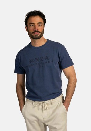 Man met donker haar en baard, draagt een marineblauw "N.Z.A. New Zealand 1923" t-shirt en beige broek met trekkoorden, handen in de zakken.