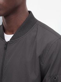 Primo piano di una persona che indossa una giacca bomber grigio scuro con zip e colletto a coste, con una camicia bianca sotto.