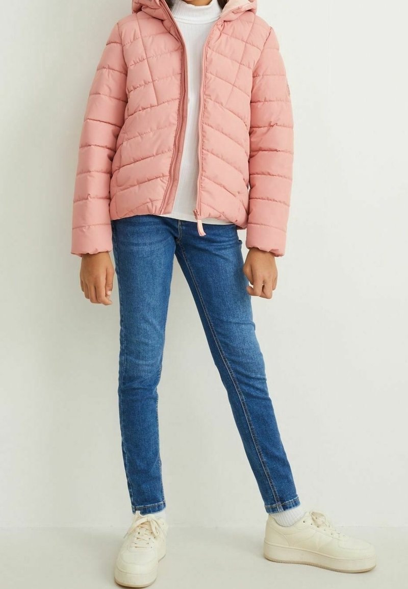 Giacca imbottita rosa con cappuccio, caratterizzata da cuciture diagonali, chiusura a zip e una vestibilità leggermente cropped, abbinata a jeans in denim blu e sneakers bianche.