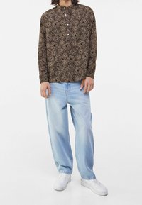 Långärmad brun skjorta med ett upprepande blommönster, kombinerad med ljusblå vida jeans och vita sneakers.