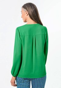 Blusa verde a maniche lunghe con scollatura rotonda, texture liscia, dettaglio plissettato sul retro e polsini leggermente arricciati. Pantaloni a motivo visibili sotto.