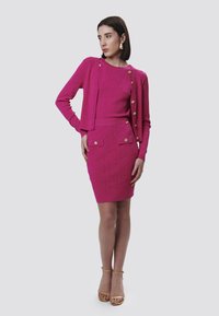 Fuchsia gestrickter Pullover und Bleistiftrock-Set mit goldenen Knöpfen und geometrischer Textur. Das Model posiert mit einer Hand auf der Hüfte und trägt Riemchensandalen.