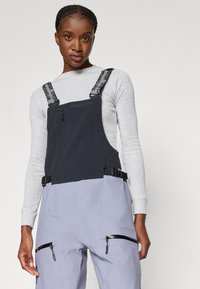 Črne in sive delovne hlače z nastavljivimi trakovi in zadrgami na žepih. Tkanina ima mat površino in sproščen kroj, primeren za nošenje na prostem.