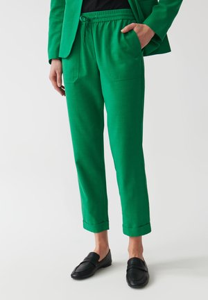 Groene, lichte broek met een elastische tailleband, twee zijzakken en opgerolde mouwen. Gecombineerd met zwarte loafers, wat een ontspannen pasvorm laat zien.
