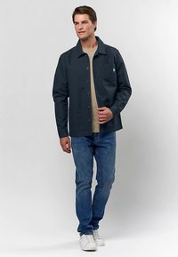 Homme portant une veste sombre boutonnée sur une chemise beige, un jean bleu et des baskets blanches, debout devant un fond clair uni.