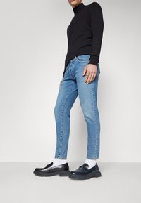 Fekete garbó, kék slim fit farmernadrág, és fekete loaferek vastag talppal. Az öltözet fehér zoknikat és gyűrűt tartalmaz a bal kézen.