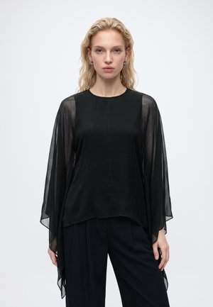 Femme aux cheveux blonds portant un haut noir avec des manches fluides transparentes et un pantalon noir, face à l’avant sur un fond uni.