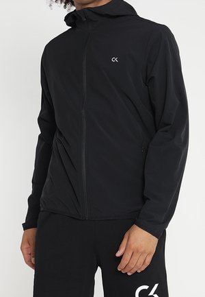 Mężczyzna w czarnej bluzie na suwak i czarnych spodniach z białymi logo "CK" na klatce piersiowej i udzie, stojący na jednolitym tle.