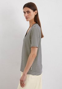 Schwarz-weißer gestreifter T-Shirt mit kurzen Ärmeln und V-Ausschnitt sowie subtilen Etikett-Details. Getragen mit einer hellen Hose, flache Stofftextur.