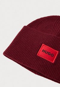 Burgundský rebrovaný pletený čiapka s prehnutým okrajom a červeným štvorcovým nášivkom s nápisom "HUGO" čiernymi písmenami.