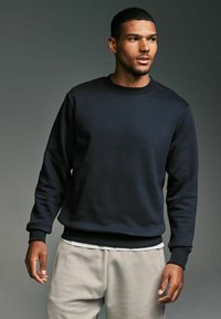 Marinblå crew-neck sweatshirt i mjukt material med ribbade ärmslut och nederkant. I kombination med ljusgrå byxor, vilket visar en avslappnad passform.