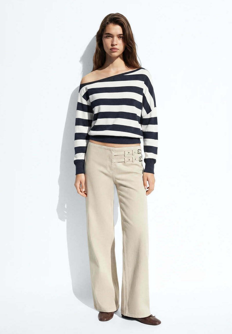 Femme portant un haut rayé noir et blanc à épaules dénudées et un pantalon beige à jambes larges avec des détails de boucles, debout devant un fond blanc.