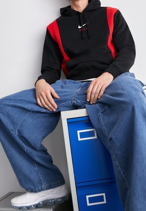 Osoba ubrana w czarną i czerwoną bluzę z kapturem Nike, z oversize'owymi niebieskimi jeansami i białymi trampkami, siedzi na niebieskiej szafce na dokumenty.