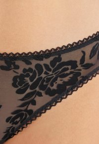 Lingerie in pizzo nero con un motivo floreale, caratterizzata da tessuto trasparente e morbide bordature ondulate per un aspetto testurizzato.