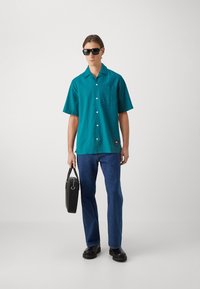 Tyrkis kortermet skjorte med teksturert mønster, frontlomme, kombinert med blå jeans og svarte tilbehør. Modellen har på seg solbriller.