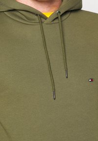 Felpa verde oliva in morbido tessuto, con tasca frontale, cordini con punte in metallo e un piccolo logo sul petto.