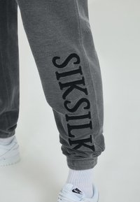 Grijze sweatpants met zwart "SIKSILK" text geborduurd op het linkerbeen. De stof lijkt zacht. Gecombineerd met witte sneakers en sokken.