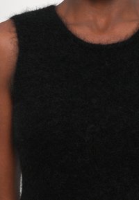 Artimas planas, kuriame matoma asmuo, vilkintis be rankovių juodą pūkuotą viršutinį drabužį, atidengiantį kaklą, petį ir viršutinę krūtinės dalį.