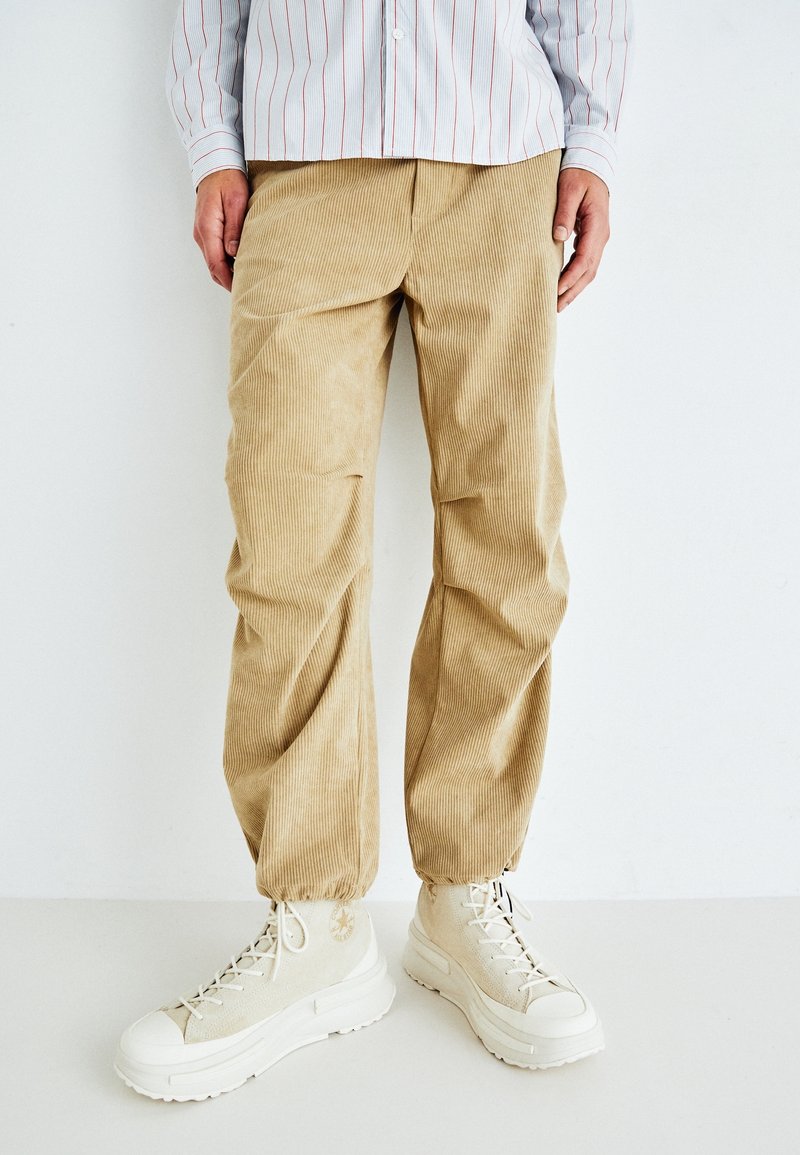 Beige corduroy broek met geribde textuur, tapered fit en elastische boorden, gecombineerd met chunky beige high-top sneakers.