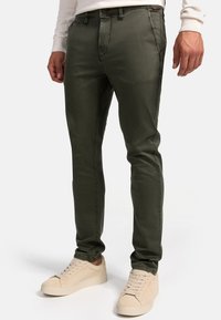 Pantalones de corte slim en verde oliva con una textura sutil, que cuentan con bolsillos frontales y cierre de botón, combinados con zapatillas de color beige claro.