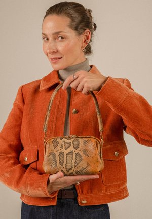 Mujer con chaqueta naranja sosteniendo con ambas manos un pequeño bolso marrón con estampado de piel de serpiente sobre un fondo neutro.