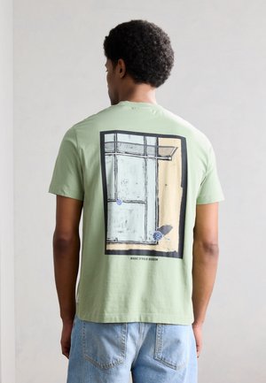 Άνδρας με κοντά σγουρά μαλλιά φοράει ανοιχτό πράσινο μπλουζάκι με αφηρημένο αρχιτεκτονικό σχέδιο και μπλε τζιν, στέκεται μπροστά σε απλό φόντο.