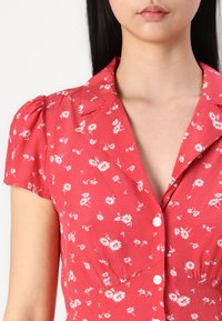 Rochie roșie cu flori, cu guler, mâneci scurte și pufoase, și modele de flori albe. Are închidere cu butoane și o textură a materialului moale.