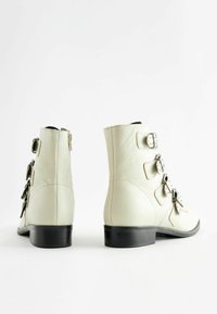 Leather ankle boots in light beige with a smooth texture, zip closure, and three decorative buckles on the outer side. Black heels.