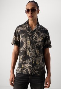 Camicia a maniche corte con bottoni di colore nero, caratterizzata da un motivo astratto grigio e giallo. Il tessuto sembra leggero e strutturato.