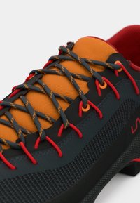 Kāpšanas apavi ar melnu un oranžu dizainu, kas aprīkoti ar teksturētām materiāliem, pelēkām šņorēm sarkaniem akcentiem un izteiksmīgu zoles rakstu.